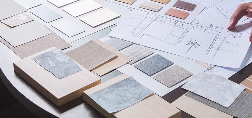 Selection of design materials and architectural sketches laid out on a table, including a variety of textured fabric and wood samples in neutral tones. The layout suggests an intricate planning process, with swatches for walls, floors, and finishes arranged meticulously alongside a hand-drawn sketch of an interior space. A hand reaches over the drawing, conveying a collaborative and hands-on approach to interior design and material selection.